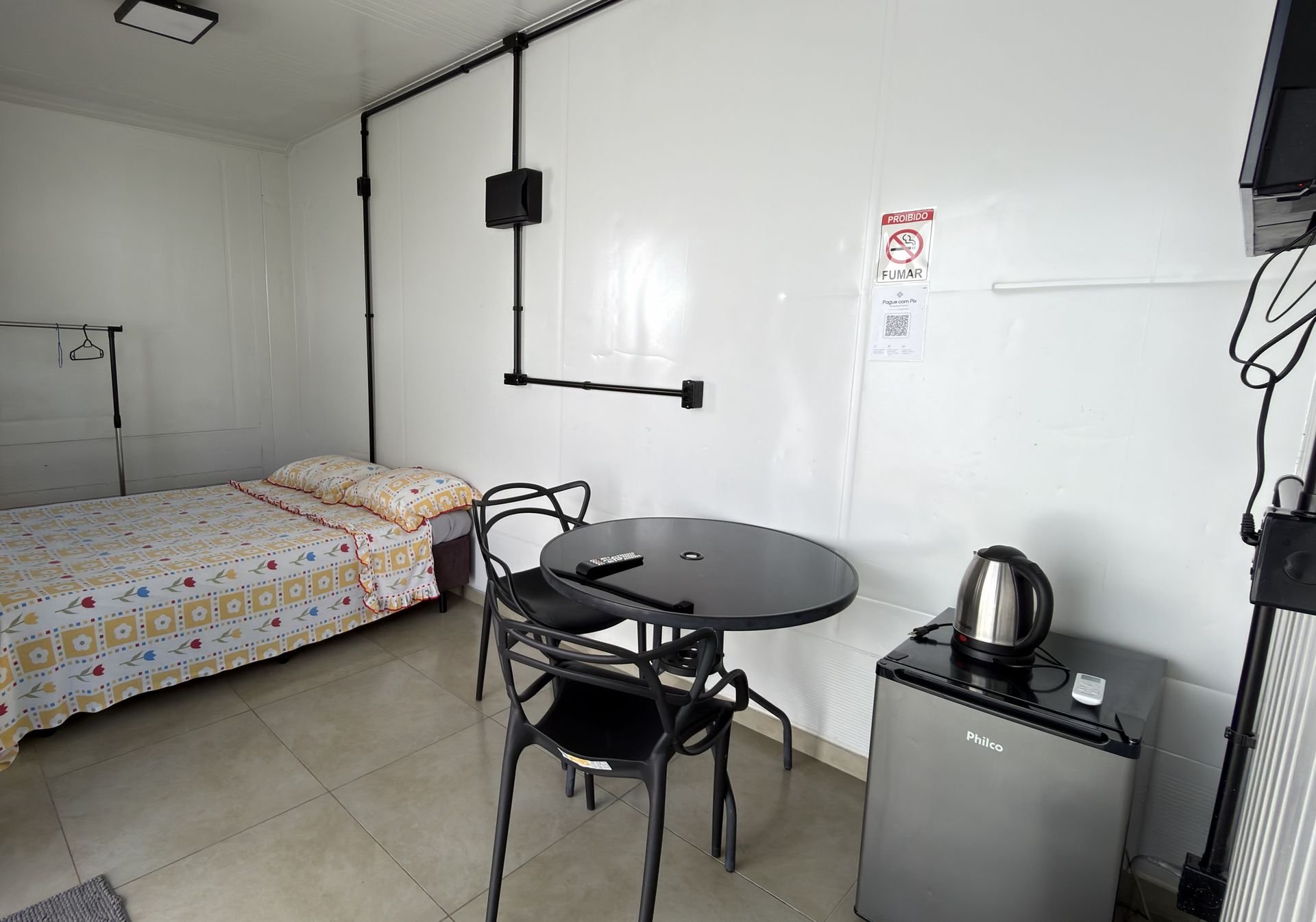 Suíte do container com cama de casal, mesinha com cadeiras, chaleira, frigobar e TV.